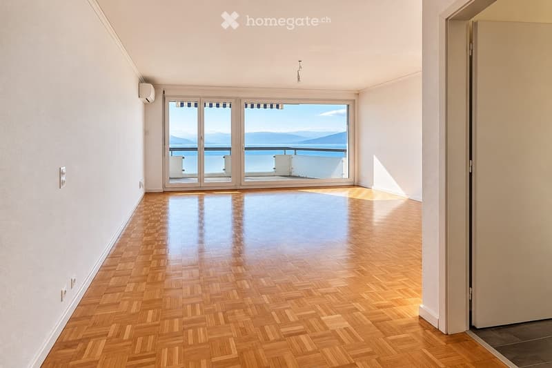 Empty room with wooden herringbone flooring, white walls, large sliding glass doors leading to a balcony with a scenic ocean view.