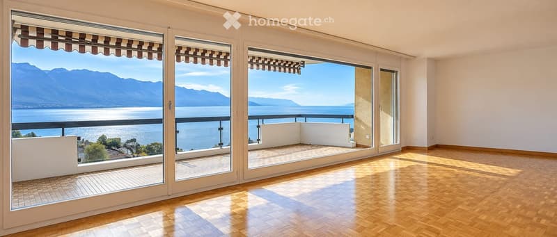 Bright, unfurnished room with large windows and sliding doors opening to a balcony overlooking a scenic lake and mountain view. The room features wooden flooring and white walls.
