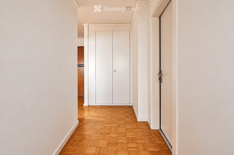 A clean, modern hallway with light-colored walls, wooden herringbone flooring, and white built-in closet doors. The space is well-lit and features a sleek, minimalist design.