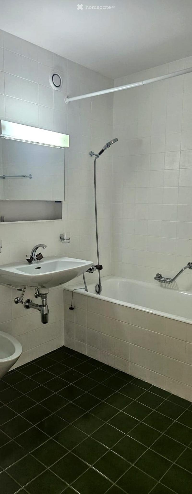 Modern bathroom with white tiles, dark green floor tiles, a sink, bathtub, showerhead, and a mirror with a built-in shelf.