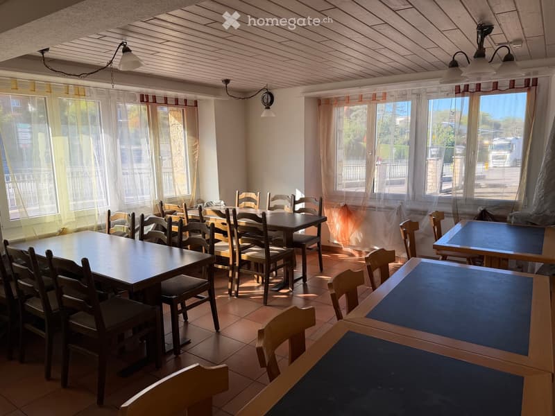Bright room with wooden tables and chairs arranged around the space, large windows with sheer curtains, and a wooden ceiling with hanging lights.