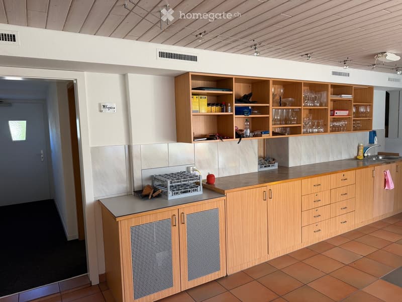 Modern kitchen with wooden cabinets, tiled floor, and open shelving displaying glassware and bottles. A dish rack and a red apple are on the counter.