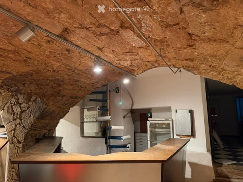 The room features a rustic, vaulted ceiling with exposed wooden beams and a stone wall. A wooden counter spans the foreground, with a mirror mounted on the wall above it. A small refrigerator and oven are visible in the background, along with several draw