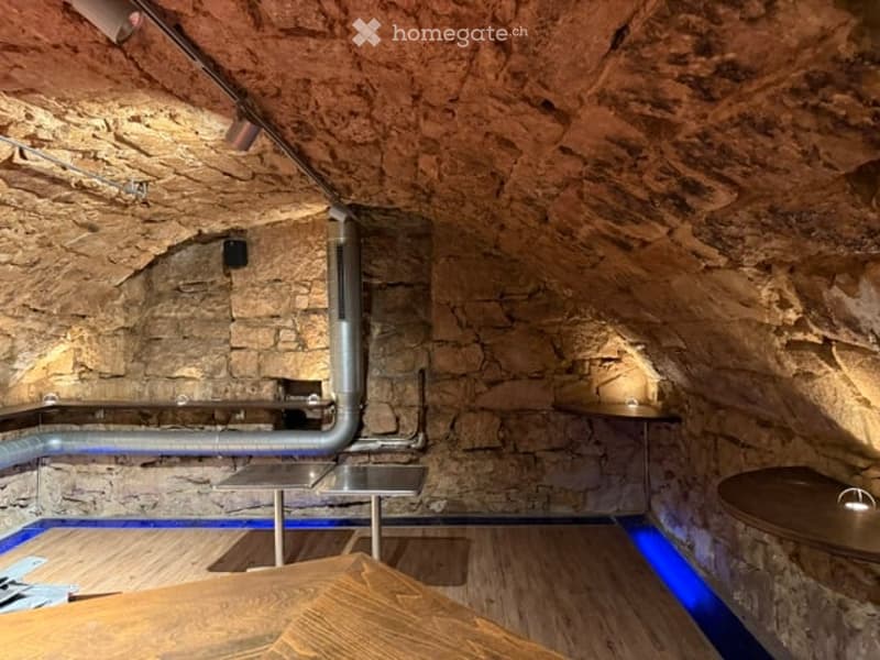 A modern, rustic-style room with exposed stone walls and wooden flooring. Features include built-in shelves, a small table, and a stool, illuminated by ambient blue lighting.