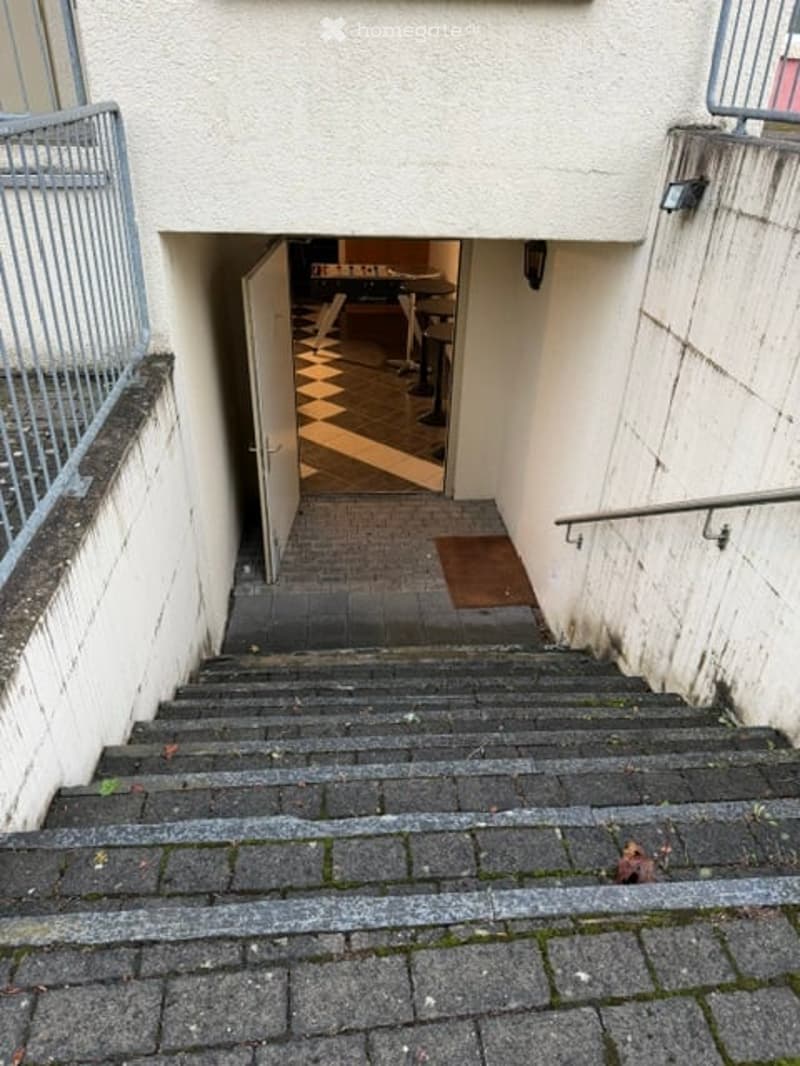 A narrow, raised entranceway leading to a room with a checkered floor, visible through an open door. The entrance is flanked by white walls and a metal railing on one side.