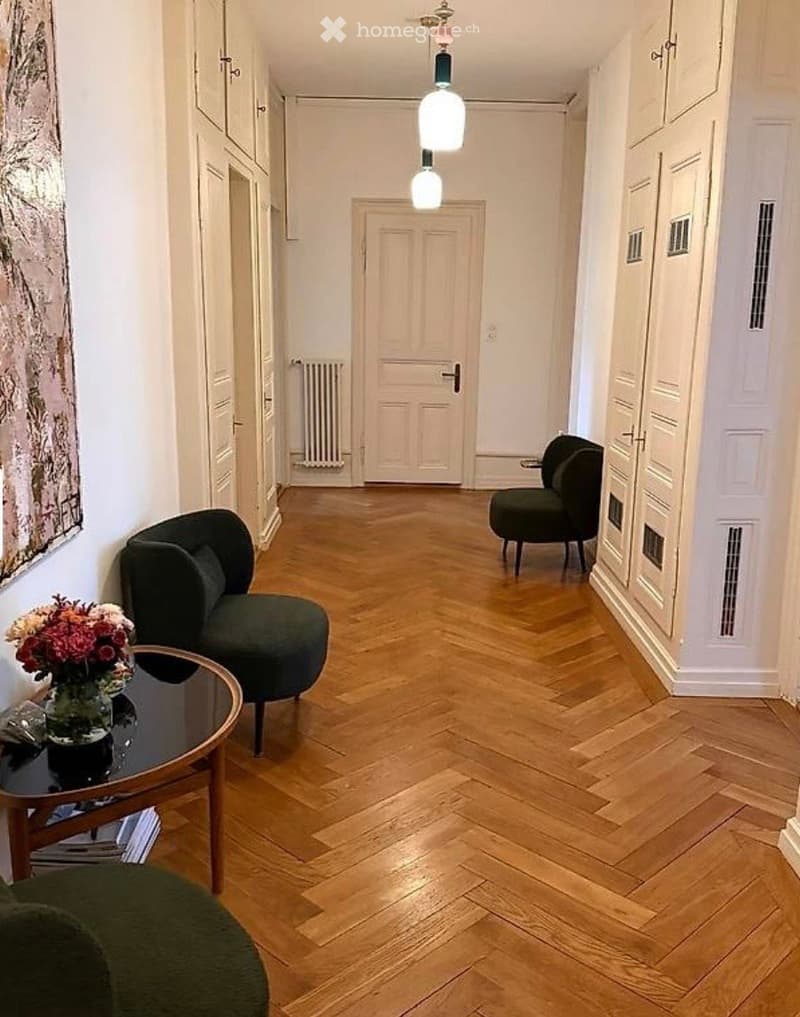 A modern hallway with herringbone wooden flooring, white walls, and two dark green chairs flanking a small round table with a vase of flowers. A closed door leads to another room.