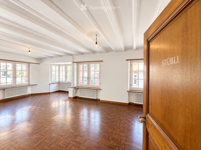 Empty room with wooden herringbone flooring, white walls, large windows, radiators, and exposed wooden beams on the ceiling.