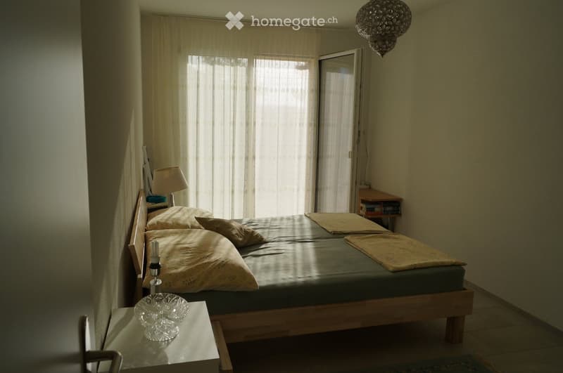 Cozy bedroom with a wooden bed, nightstand, lamp, and a small table with a decorative bowl. Light-colored curtains cover the window, and a chandelier hangs from the ceiling.