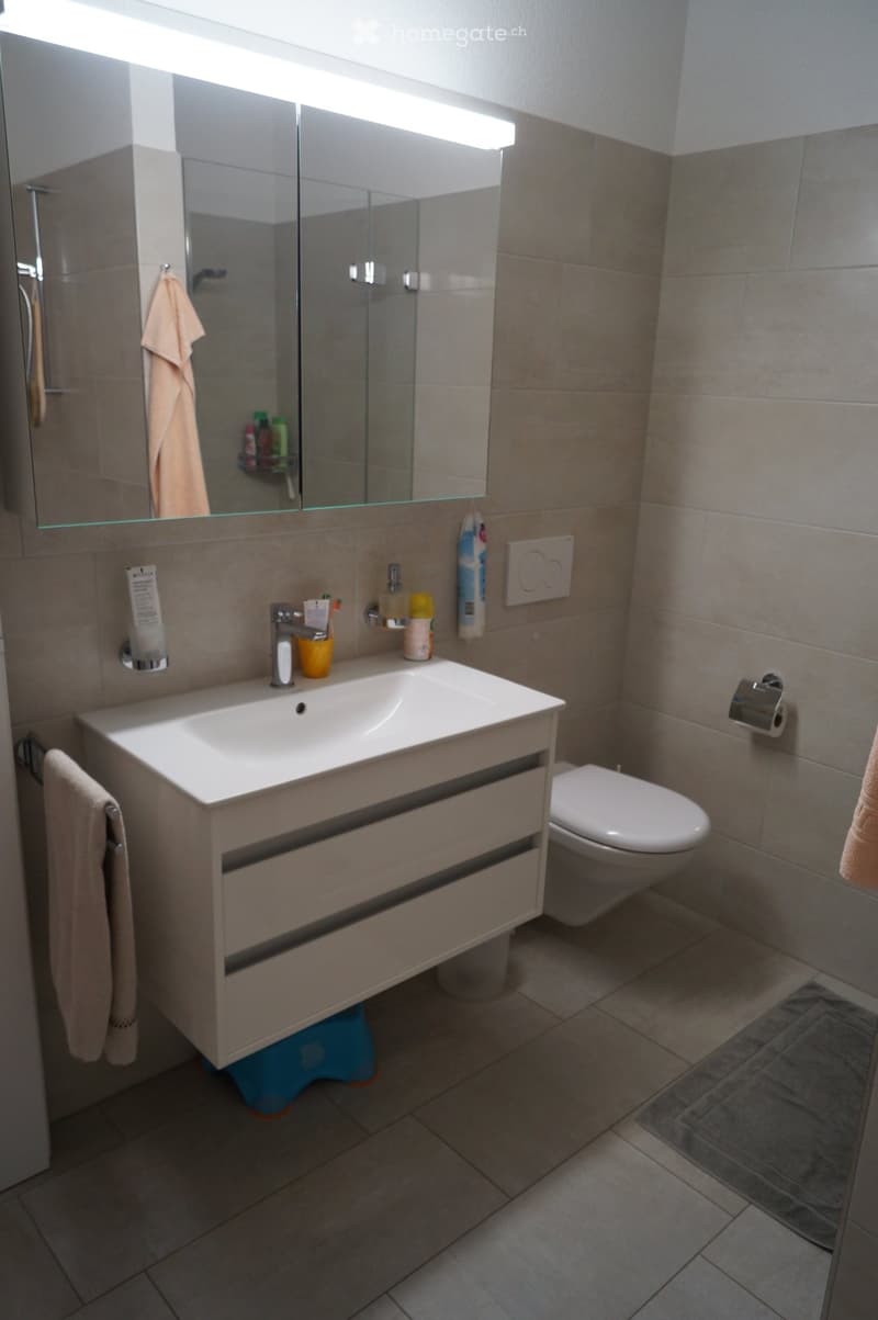 Modern bathroom with a white sink, mirror, and toilet. Beige tiles and a light-colored cabinet are visible.