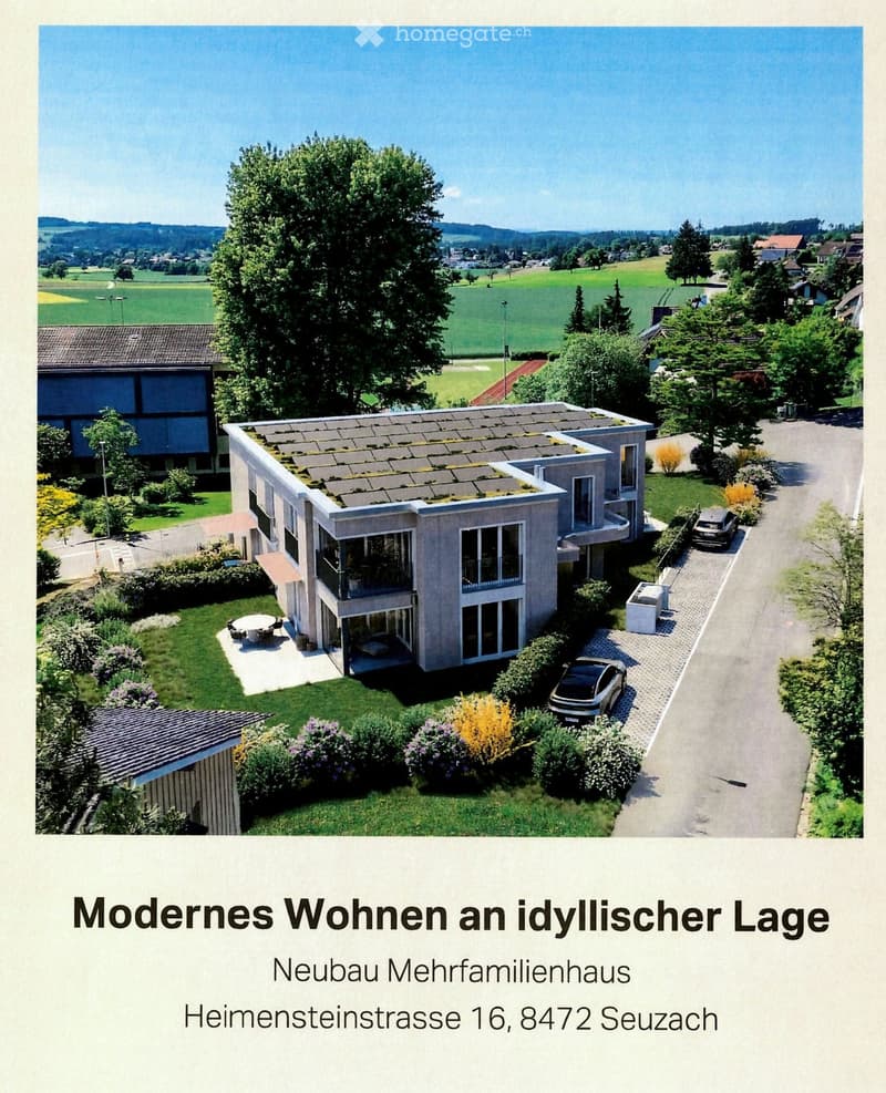 A modern two-story house with a flat roof, large windows, and a balcony. The property includes a well-maintained garden, a parking area, and a scenic rural backdrop with fields and trees.