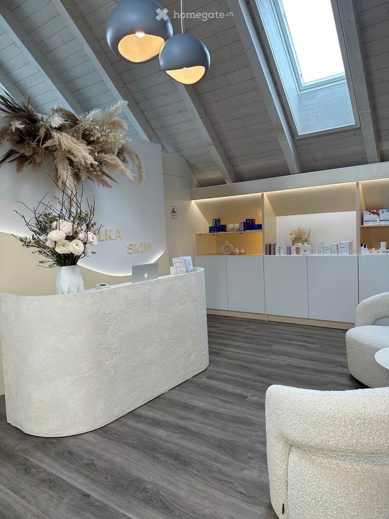 Modern reception area with a sleek white reception desk, laptop, vase with dried flowers, shelves with skincare products, and seating area with a round stool and small table.