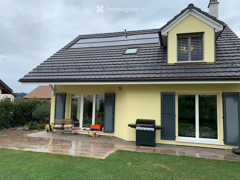 A modern single-story house with a yellow exterior, dark grey shutters, and a tiled roof. The patio area features a wooden bench, a black grill, and a toy car, with a well-maintained lawn in the foreground.