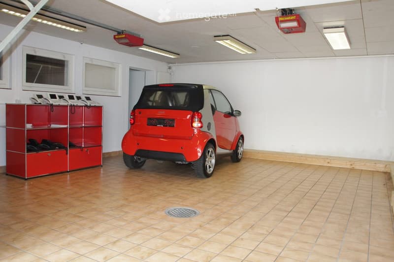 {"type": "Garage", "objects": ["car", "storage unit"], "description": "Clean, well-lit garage with tiled floor, white walls, and a red Smart car parked inside. A red storage unit with multiple compartments is visible on the left side.", "localized_descrip