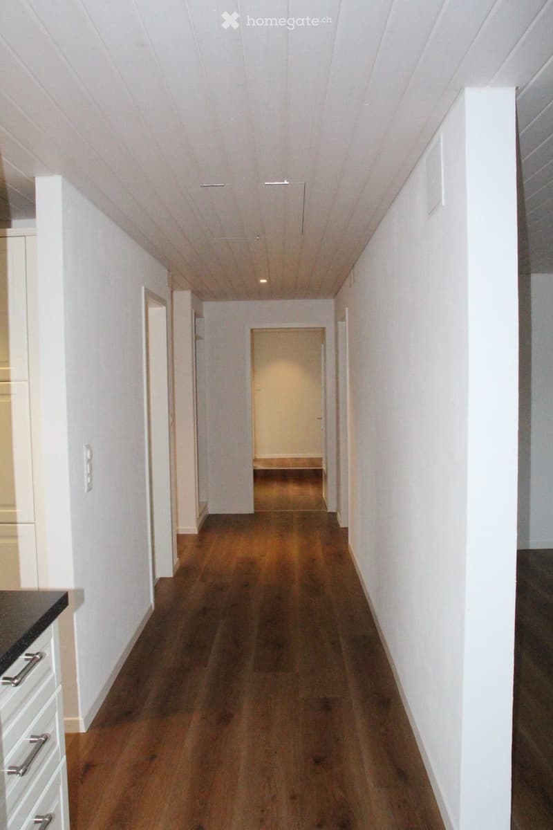 A long, narrow hallway with white walls and wooden flooring, featuring several doors and a visible kitchen area on the left side.