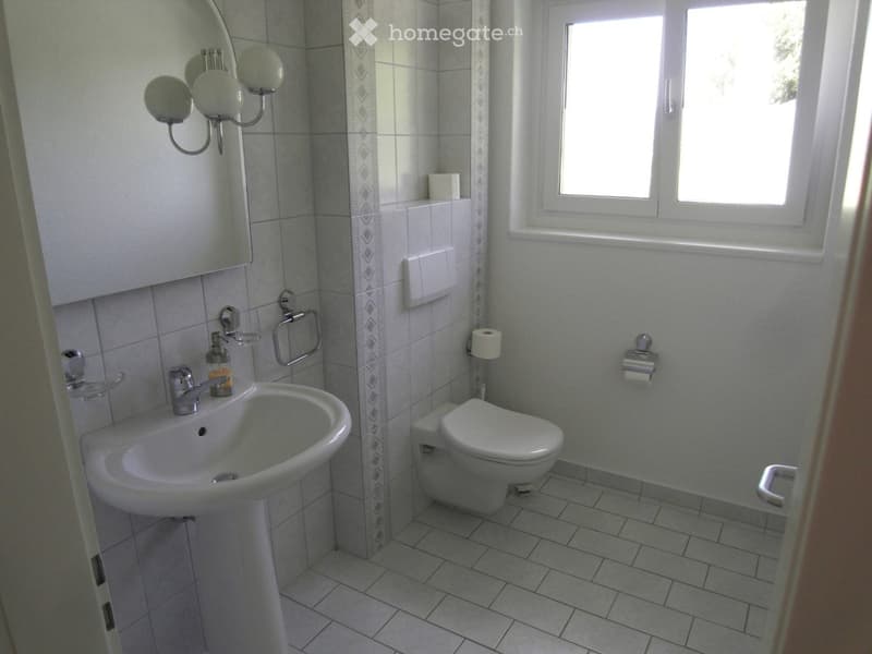 A small, modern bathroom with a white sink, toilet, and mirror. The walls are tiled with a decorative border, and there is a window providing natural light.