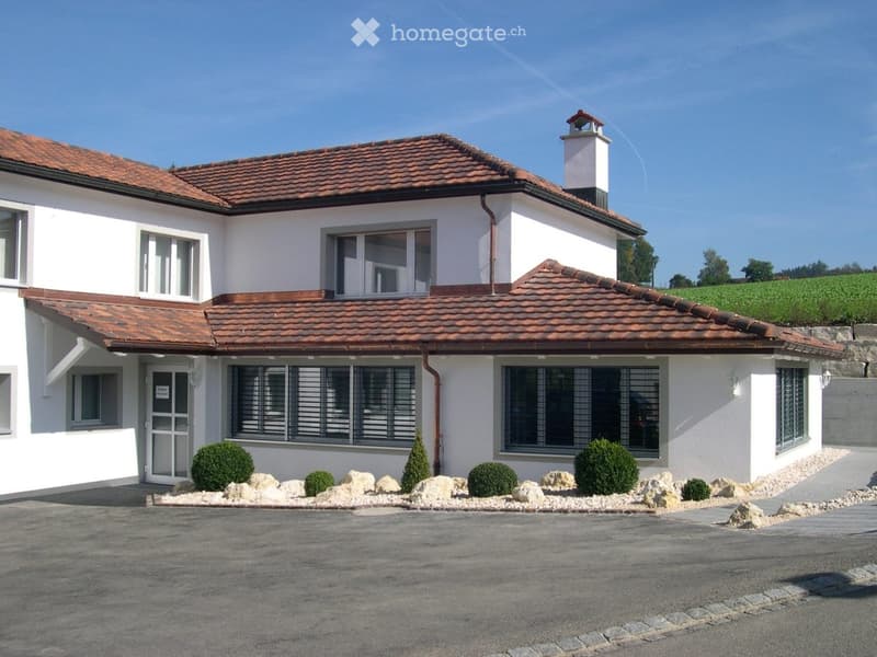Two-story white house with red-tiled roof, multiple windows, and a small chimney. The house is surrounded by a landscaped area with small shrubs and rocks.