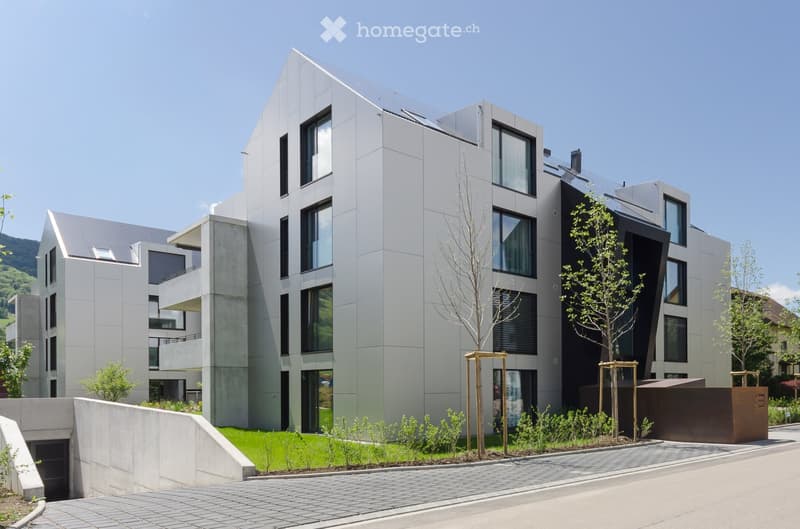 Modern multi-story residential building with sleek, contemporary design featuring large windows, concrete and steel elements, and landscaped surroundings with trees and shrubs.