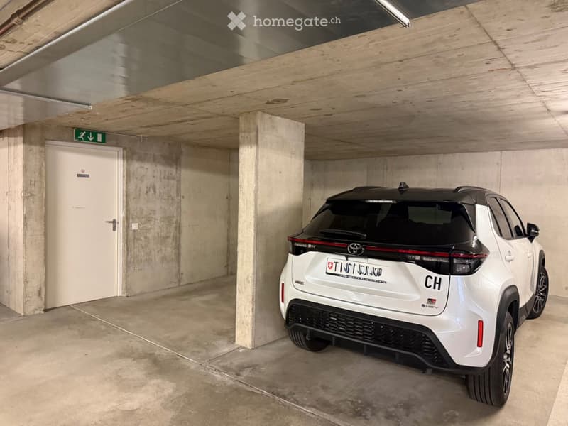Underground parking area with a white SUV parked inside, featuring concrete pillars, a white door, and a green directional sign on the ceiling.