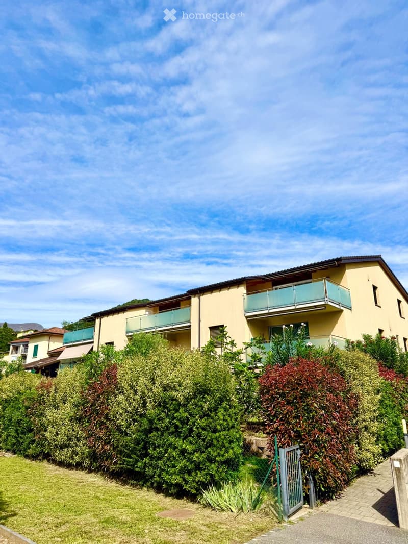 A two-story residential building with light yellow walls, multiple balconies, and surrounding greenery.