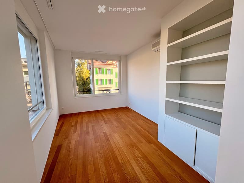 Empty room with wooden flooring, white walls, built-in shelving unit, and large windows offering a view of the street.