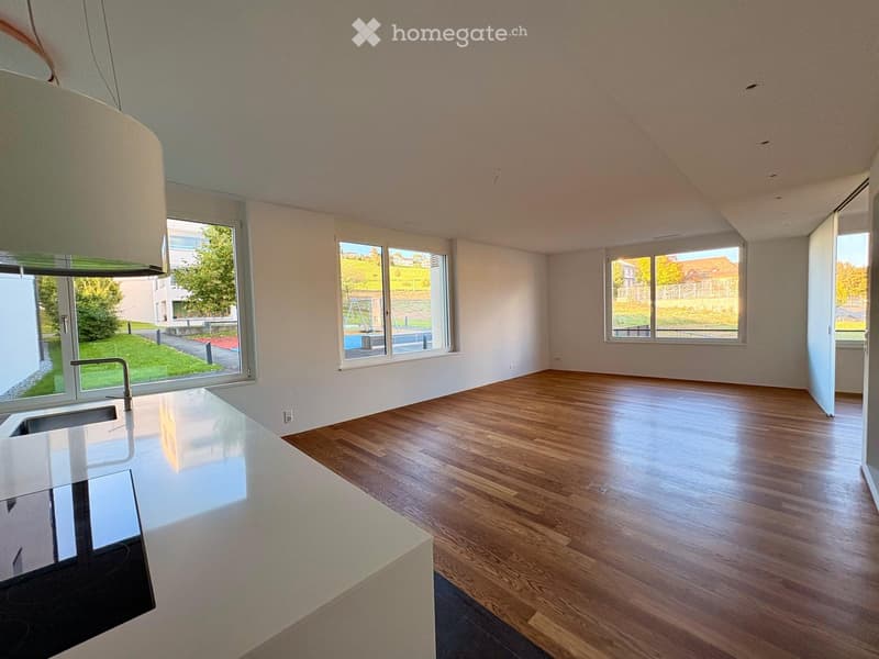 Empty kitchen with wooden flooring, white walls, large windows, and a modern kitchen island with a sink.