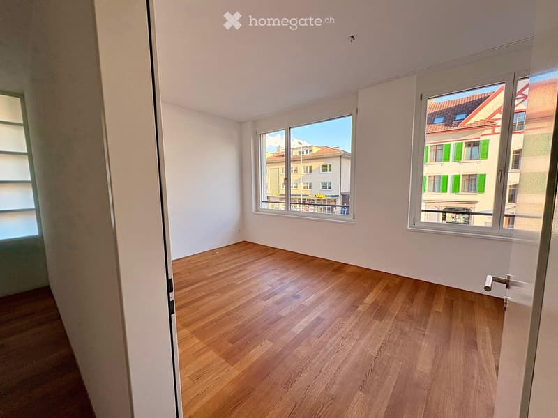 Empty room with wooden flooring and large windows showing a view of neighboring buildings.