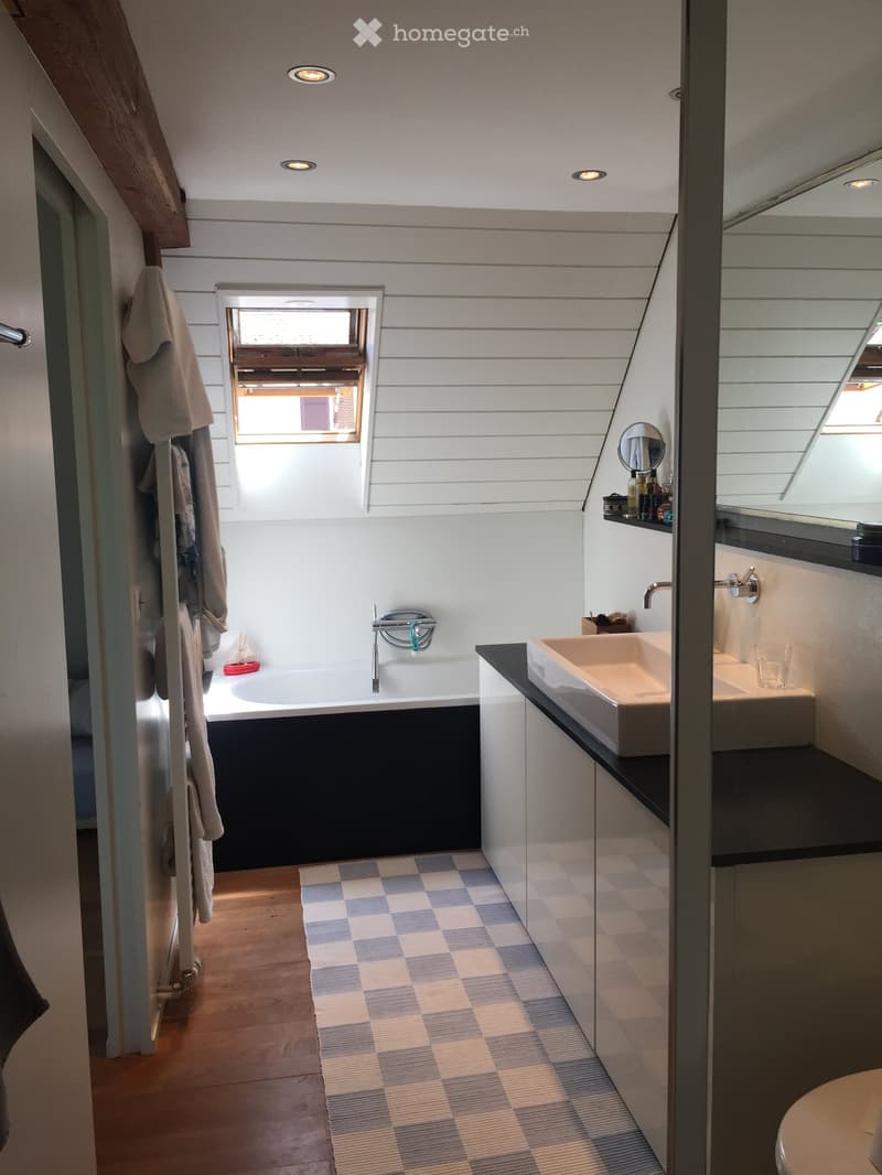 Modern bathroom with a bathtub, double sink, mirror, and a window. The space features wooden flooring and a checkered rug.