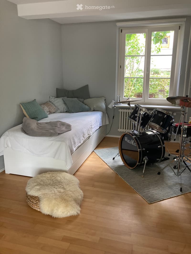 Cozy bedroom with a white bed, grey pillows, a drum set, a fluffy pet bed, and a grey rug near the window.