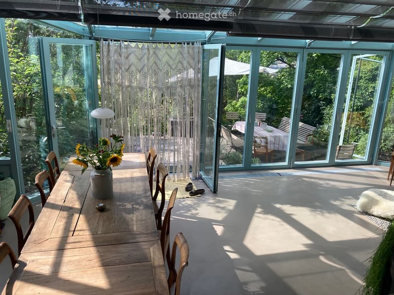 Bright, airy room with large glass walls and doors leading to a garden, featuring a wooden dining table with chairs, a vase of sunflowers, a white hanging light fixture, and a fluffy white rug on the floor.