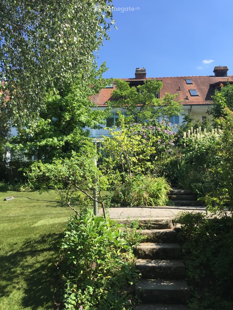 Garden with lush greenery, stone steps leading up to a house with a red-tiled roof and white walls.