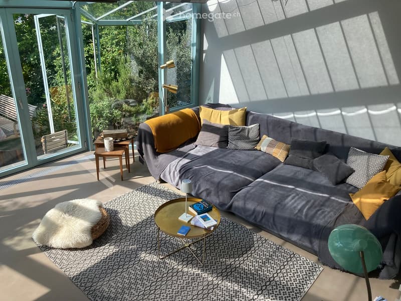 Modern living room with large windows, grey sectional sofa, colorful pillows, coffee table, side table, floor lamp, and books.