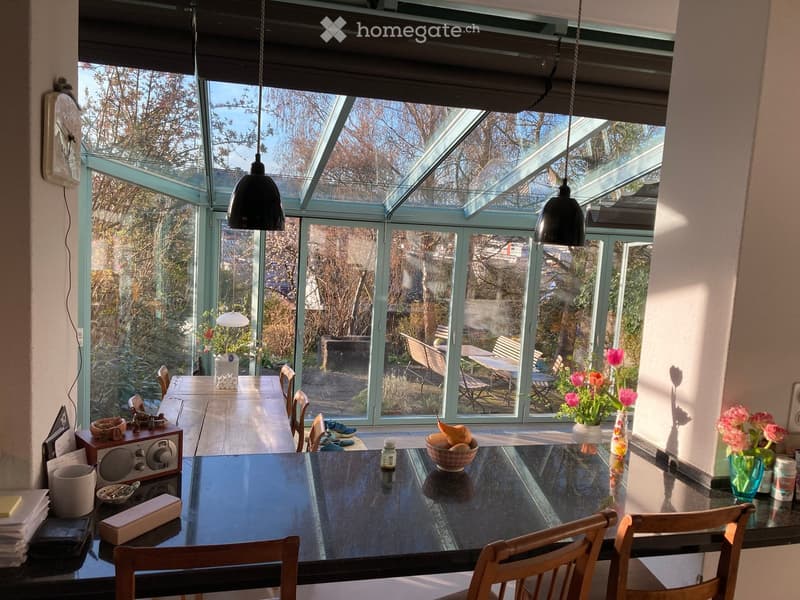 Bright, airy dining area with large glass walls and a skylight, featuring a black dining table with chairs, vases with flowers, and a bowl of fruit on the table.