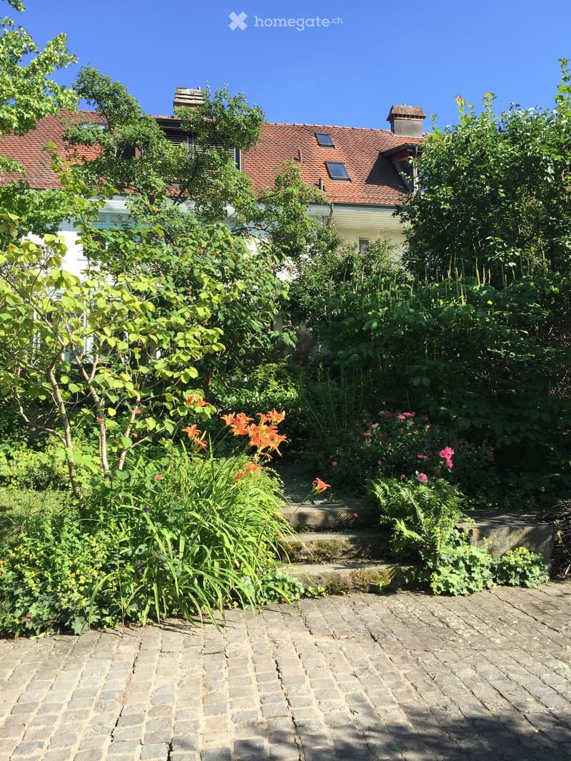 A well-maintained garden with a stone-paved pathway, surrounded by lush greenery and colorful flowers, with a house partially visible in the background.