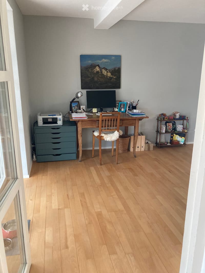 A well-lit study or home office with wooden flooring, a wooden desk, a chair, a printer, a screen, a lamp, a shelf with various items, and a painting on the wall.