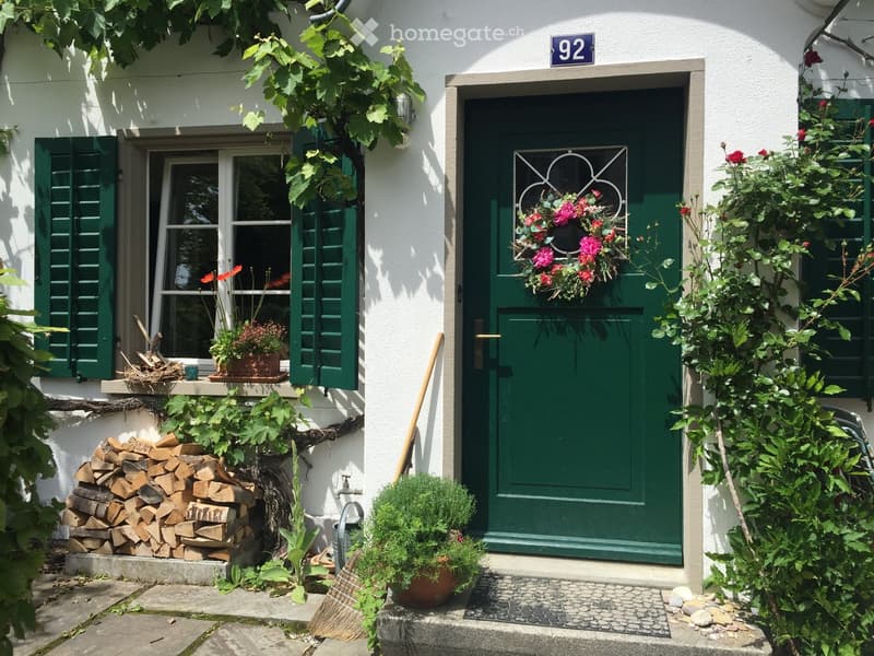 Front entrance of a house with a green door, floral wreath, potted plants, and a stack of firewood.