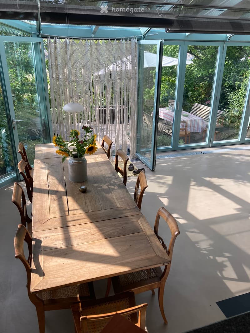 Bright, airy room with large glass walls and a wooden dining table surrounded by chairs. A vase with sunflowers sits on the table, and a hanging lamp provides light. The room has a view of a garden through the glass walls.