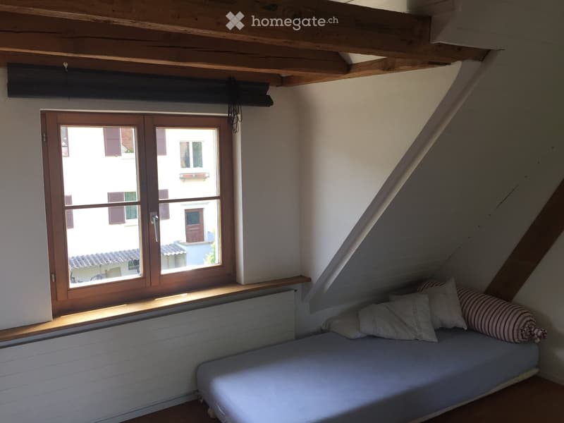 Cozy attic bedroom with a single bed, pillows, wooden window frame, and slanted wooden ceiling beams.