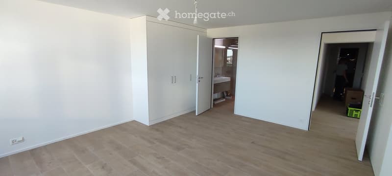 Empty room with light-colored walls, wooden flooring, and built-in white closet. Open doors lead to a bathroom and another room with a green basket visible.