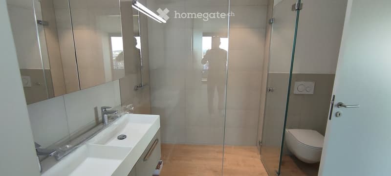 Modern bathroom with a double sink, large mirror, glass-enclosed shower, and a toilet. The space features light-colored tiles and a sleek design.