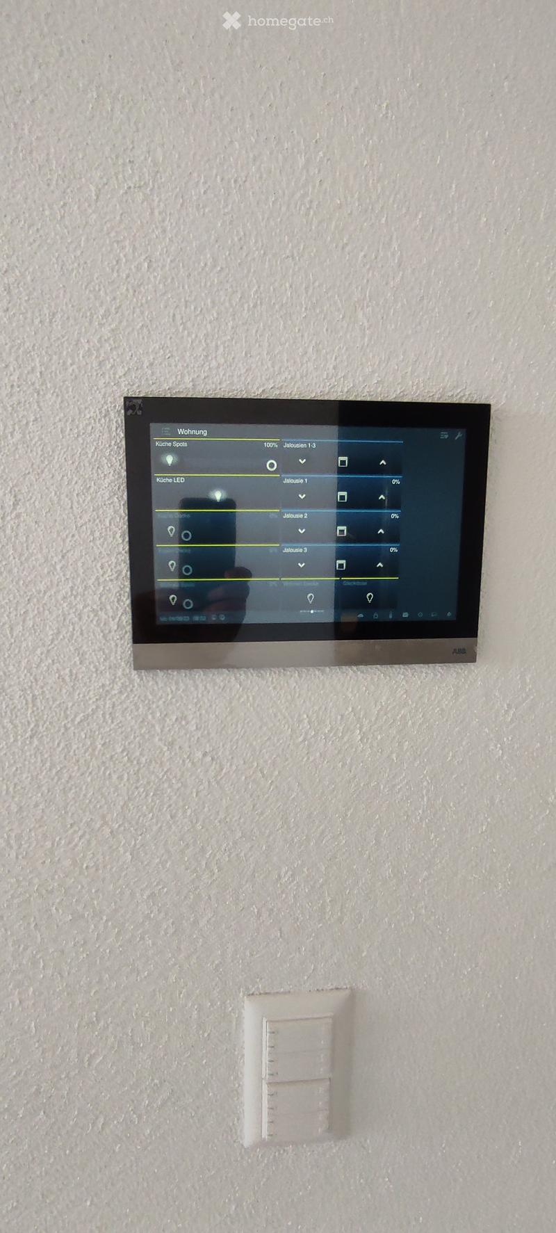 A modern wall-mounted control panel with a touchscreen display and a smaller switch below it, mounted on a textured white wall.