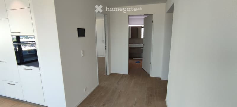 Empty hallway with wooden flooring, white walls, and open doors leading to other rooms. A small screen is mounted on one wall, and a cabinet is visible on the left side.