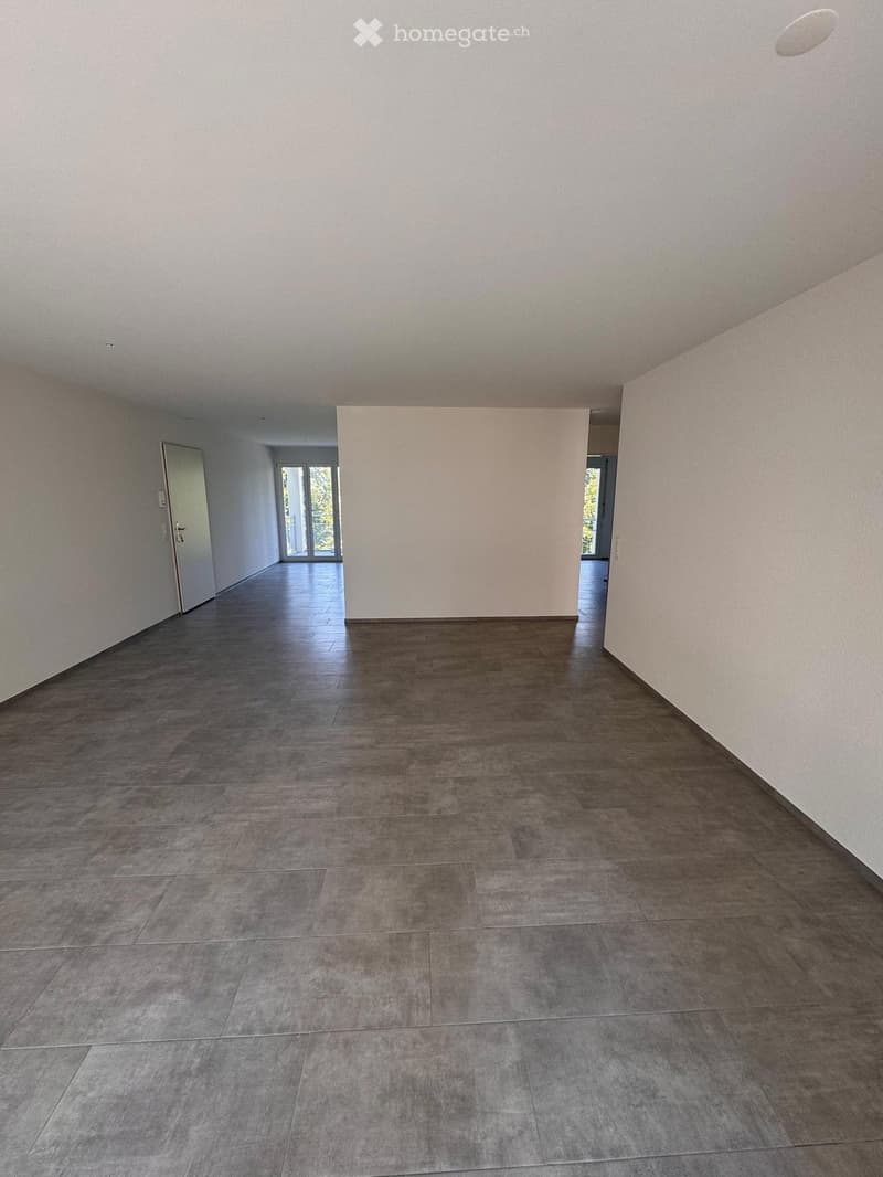 Empty hallway with grey tiled floor and white walls, featuring a closed door on the left and an open doorway leading to another room at the end.
