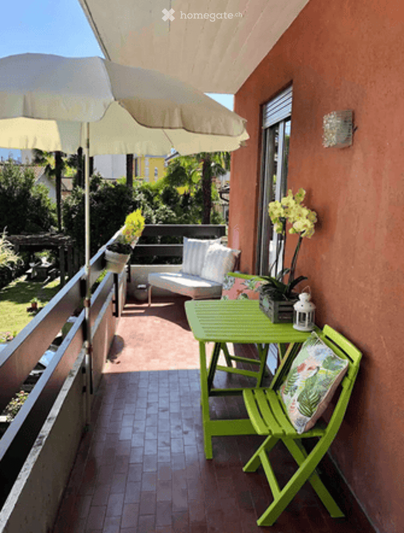 Balcony with green folding table and chairs, white umbrella, potted plants, and a view of a garden.