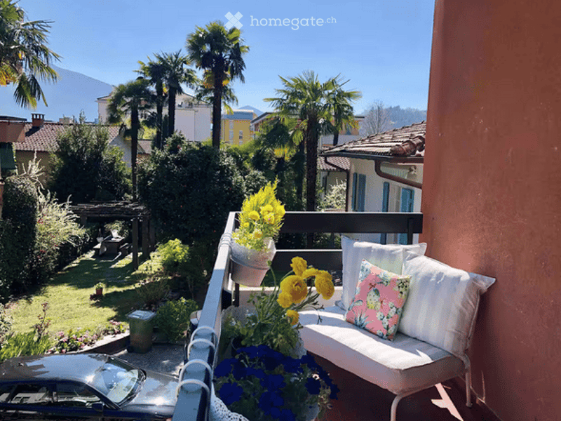Balcony with a white bench, colorful cushions, and potted flowers, overlooking a garden with palm trees and a distant mountain view.