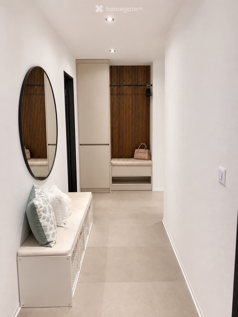 Modern hallway with white walls, beige tiled floor, built-in bench seating on both sides, decorative pillows, a large oval mirror, and a coat closet with a hanging coat and a handbag on the bench.