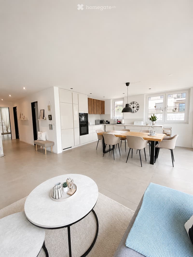 Modern open-plan living and dining area with white walls, light wood flooring, and large windows. Features a round coffee table with decorative items, a sofa, a dining table with chairs, a bench, and a kitchen area with cabinets, an oven, and a microwave.