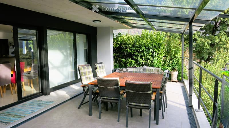 A modern, covered balcony with a wooden dining table and chairs, surrounded by lush greenery and a view of the garden.