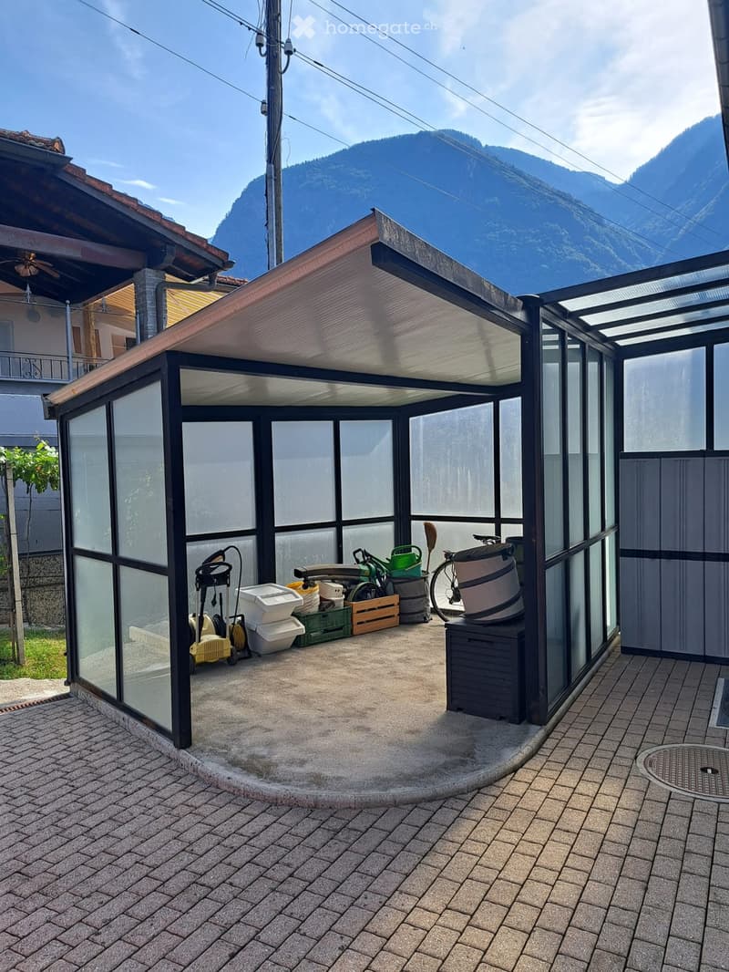 A covered outdoor area with a concrete floor, glass walls, and a variety of stored items including a bicycle, garden tools, and storage boxes.