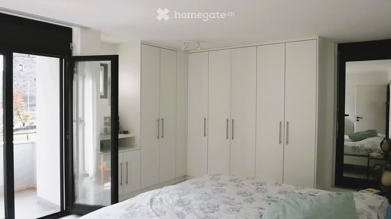 Modern bedroom with a large bed, white built-in wardrobes, a large mirror, and a nightstand. The room features large windows and a sliding glass door leading to a balcony.