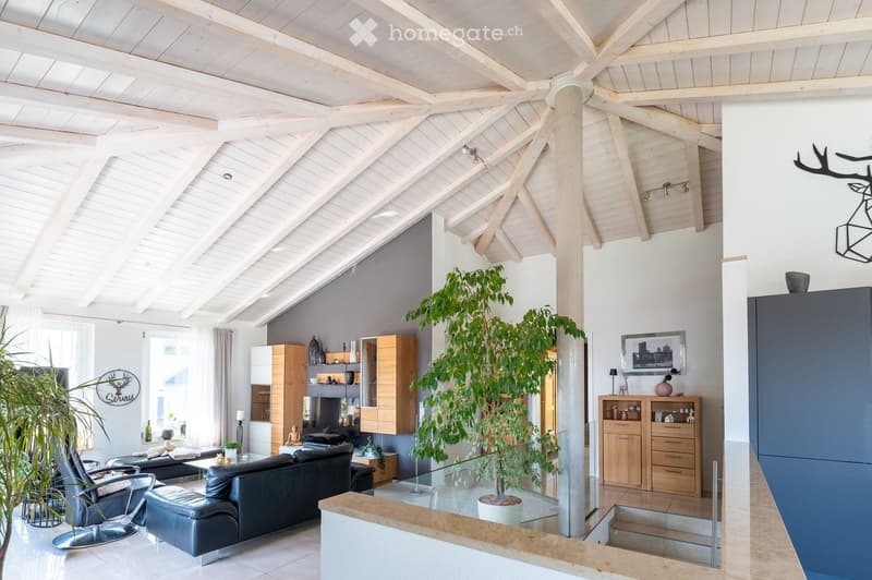 Modern living room with exposed wooden beams, white walls, and large windows. Features a black leather sofa, a glass coffee table, a potted plant, and wooden cabinets.